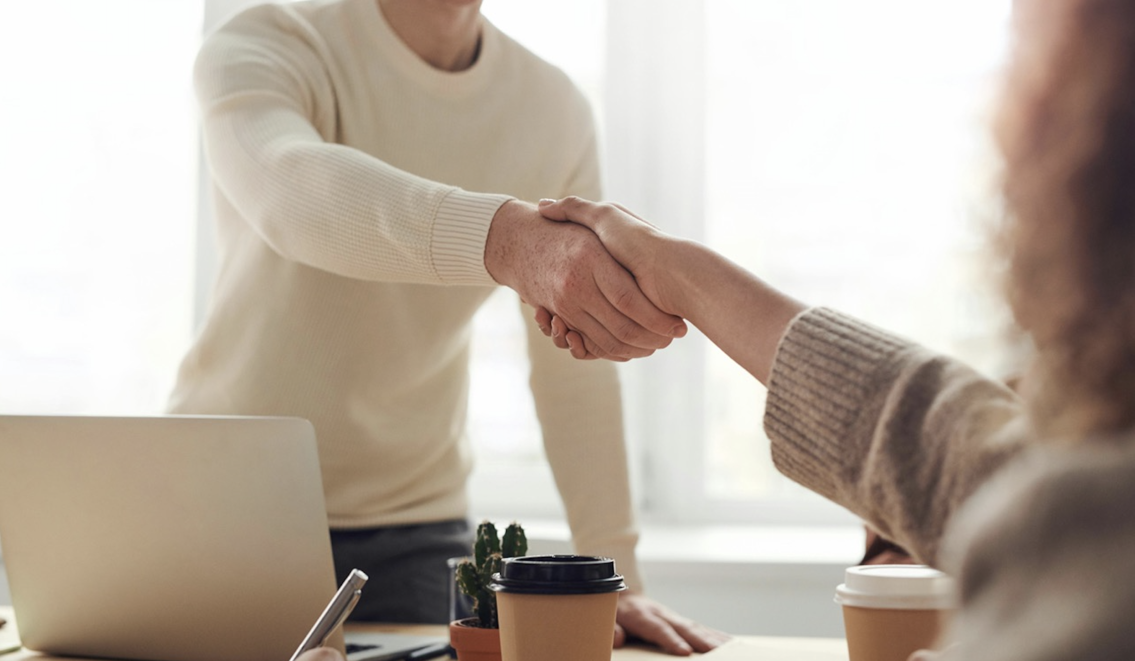 Close-up of a handshake between two professionals in a bright, modern office setting over a desk featuring coffee cups and a laptop.